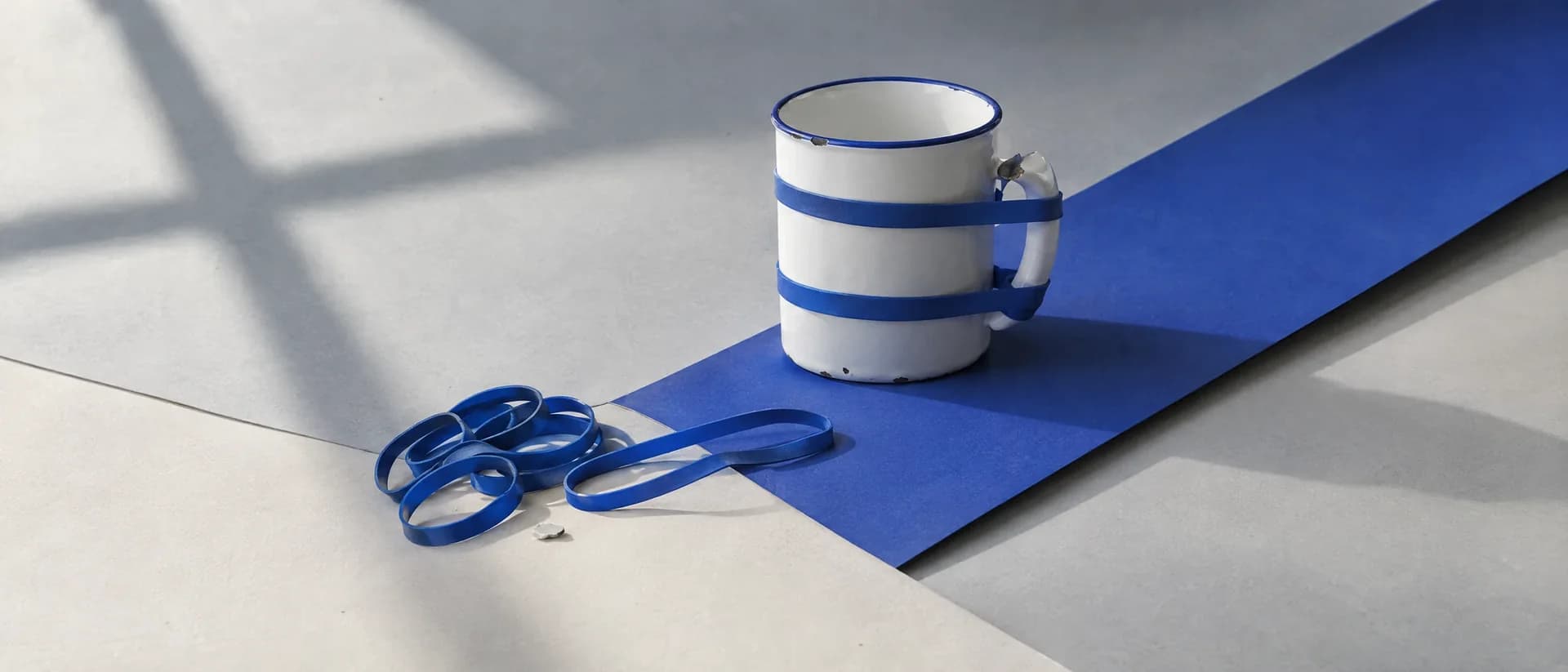 An enamel mug with a snapped handle held on by cobalt rubber bands.